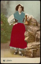 Woman in red silk apron and blue silk shawl holding a decanter.
