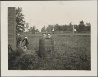 Two men, one in a barrel and one behind in field