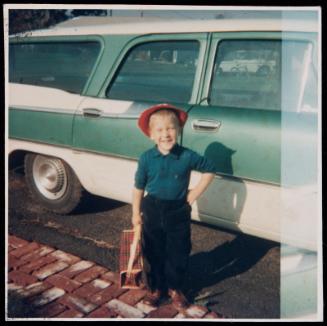 Young boy with red hat and plaid bag standing in front of car