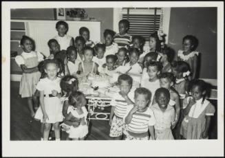 Large group of children surrounding table with birthday cake and presents