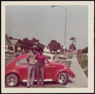 Portrait of a couple standing in front of a red VW Beetle on suburban street with palm trees