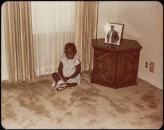 Small girl seated on the floor next to low table with framed portrait displayed on it