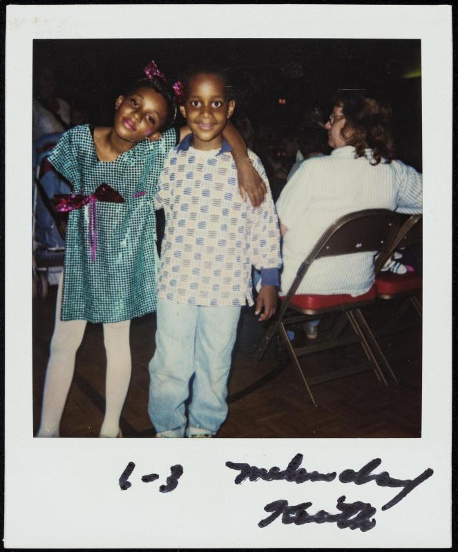Two young girls with arms around each other's shoulders next to seated figure
