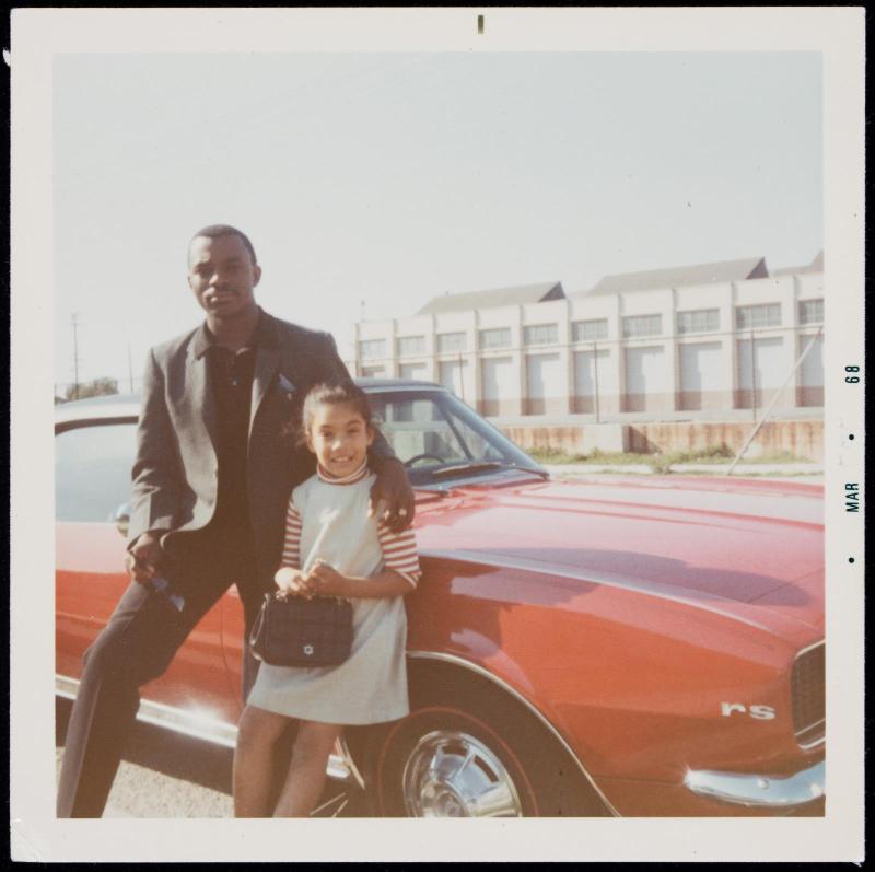 Portrait of a man and girl posed next to red car with large building behind them