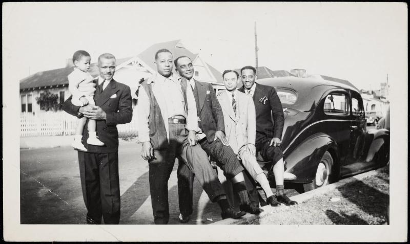 Group of five men and one child posed in front of car with feet on curb