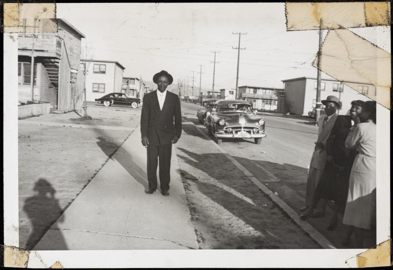 Man posed on sidewalk with shadow of the photographer, cars behind, and group watching at right