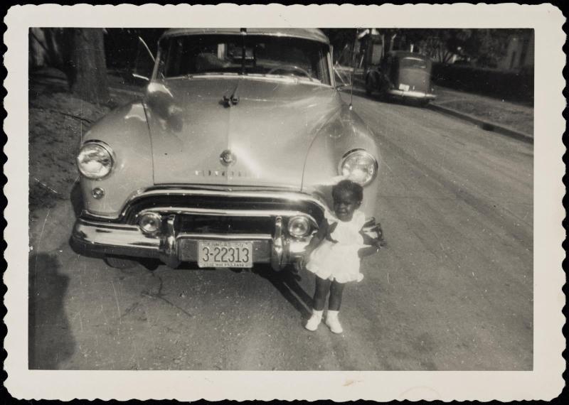 Small girl in white dress standing in front of large car