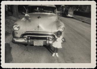 Small girl in white dress standing in front of large car