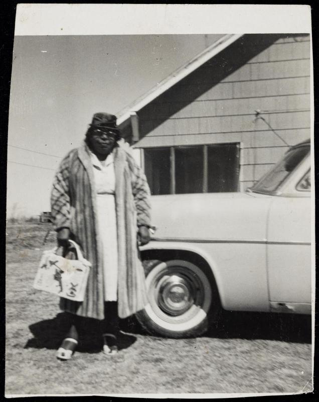 Portrait of woman standing next to car wearing glasses and fur coat and carrying large handbag