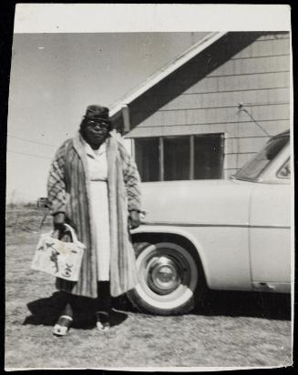 Portrait of woman standing next to car wearing glasses and fur coat and carrying large handbag