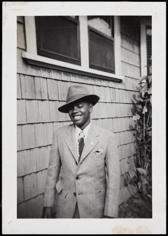 Boy wearing suit, tie, and hat in front of clapboard house