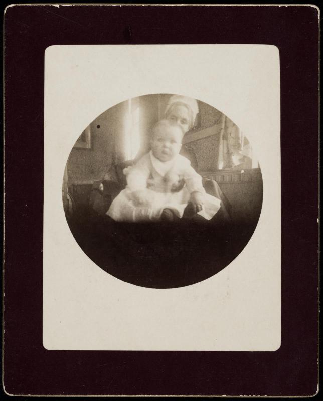 Seated baby being supported from behind by a woman mostly hidden