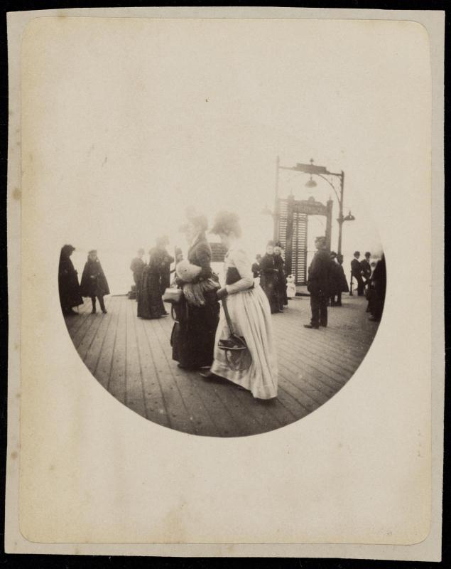 Two women walking in long skirts and hats on crowded pier or boardwalk