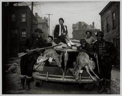 Hunters gathered around car with two dead deer lying on hood and woman seated on roof