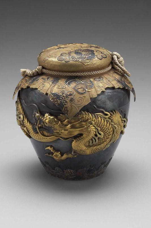 Tiered jar and cover with simulated silk brocade wrapper with mandarin duck and floral designs, tied with a real silk cord, the sides with a dragon above formal seigaiha and standing lappet ornament overlaid with sprigs of chrysanthemum